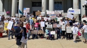 A photo of a man in a jacket holds a microphone as he speaks in front of a crowd of protesters holding signs. Signs read: "FIRE: Cordelli, Osborne, Ammon" and "OMG GOP WTF?" and "Fair Budget Now"