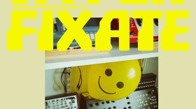 Album cover of Chad McKinney's album "Hyperfixate (Waiting for Something to Happen)," featuring an off-white color, yellow typeface with the album title, and a picture of electronic instruments in his studio along with a yellow smiley-faced balloon.