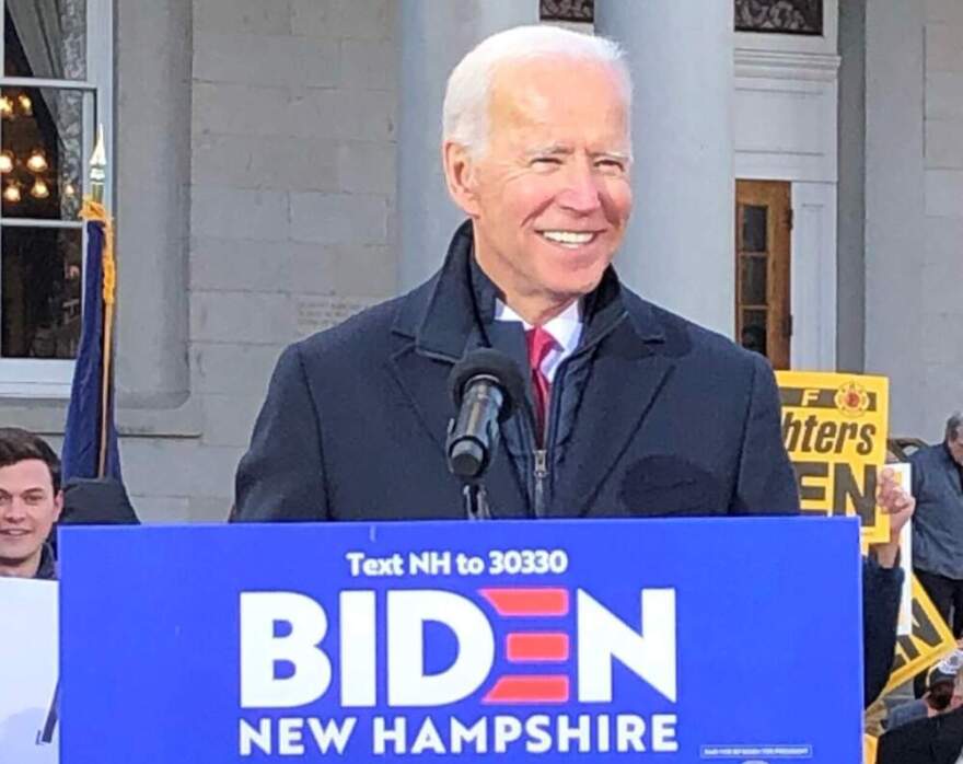 A man with white hair, a navy jacket and red tie, President Joe Biden, stands behind a sign that reads Biden New Hampshire