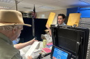 An older man wearing a tan fedora holds a long slip of paper. A younger man standing across a desk from him holds up two large envelopes of election related documents.