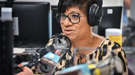 A woman sits inside a radio studio wearing a headset and talking while gesturing with her right hand.