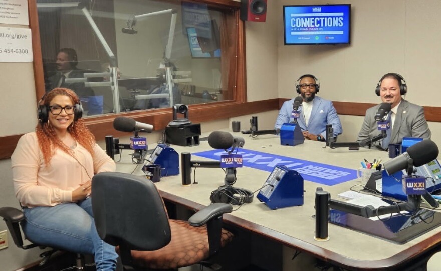 Three smiling people wearing headphones sit at a table in a radio talk studio: a woman front left has long, red, wavy hair and is wearing glasses, gold hoop earrings, a long sleeved peach top, jeans, and tan sandals; a man back left has short black hair and is wearing glasses and a white shirt under a light blue suit coat; a man back right has short black hair and a mustache and is wearing a white collared shirt and blue tie under a grey suit coat