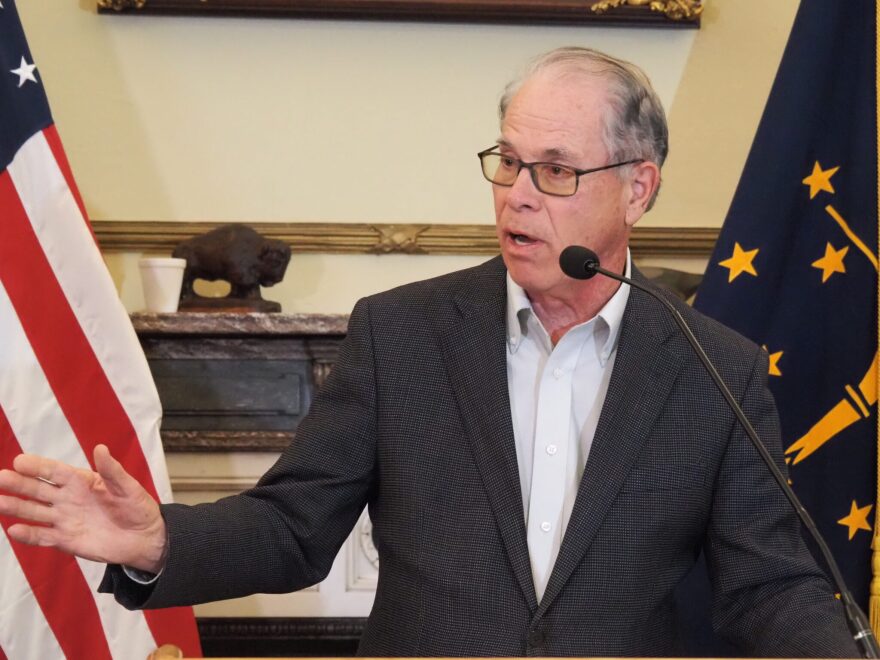 Mike Braun is wearing a blazer, standing at a microphone, speaking to reporters