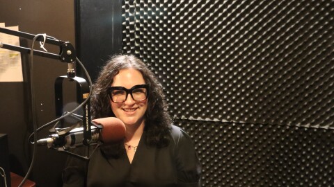 A woman wearing glasses and a black blouse sits smiling in a recording studio beside a microphone.