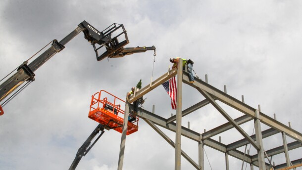 A crane lifts a beam to the top of a metal framework for a building. Two men in hardhats pull the beam over and secure it. 