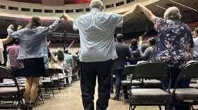 A photo of people holding hands and raising their arms, at the back of a crowd of worshippers on a large convention center floor.