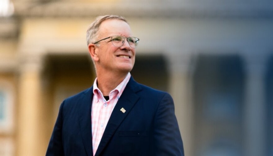 A man is standing in front of a blurred-out background. He has glasses and is wearing a dark suit and staring to his right.