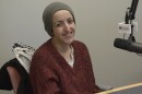 Young person wearing beanie and a red sweater seated smiling next to a microphone. 