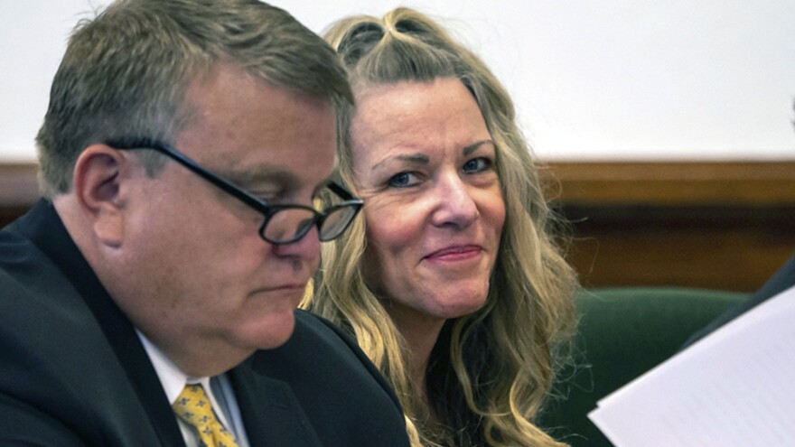 A woman looking into the camera with blonde hair. There is a man wearing glasses in front of her looking down at something on a desk. 