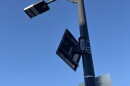A security camera is affixed to a light pole. The photo is a worm's eye view. The camera has a very small solar panel above it, also affixed to the pole. 