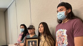 Four relatives of Irma and Joe Garcia pose for a photo. A woman holds a sign that says "You can't wash the blood of the innocent off your hands," while another woman and a teenager hold a photo of the teacher and her husband. A young man stands next to them.