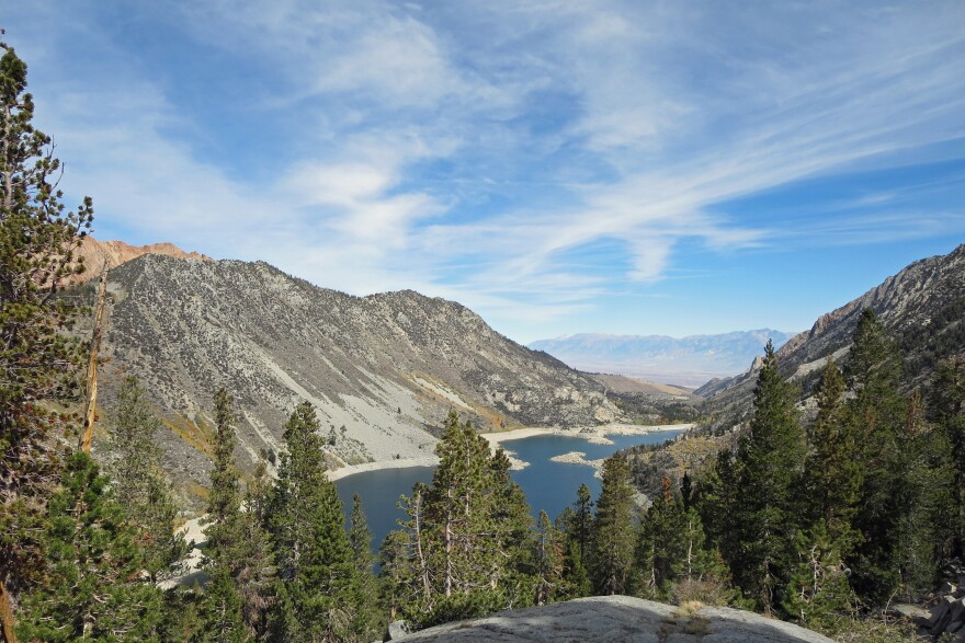 A lake sits between two rocky mountains. In the foreground are large conifer trees.