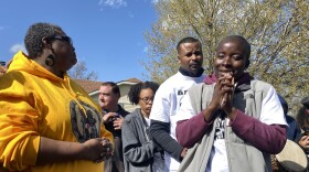 A woman in a yellow hoodie stands and looks at a woman in a purple hoodie with a grey vest over it. Behind them are people looking solemn.