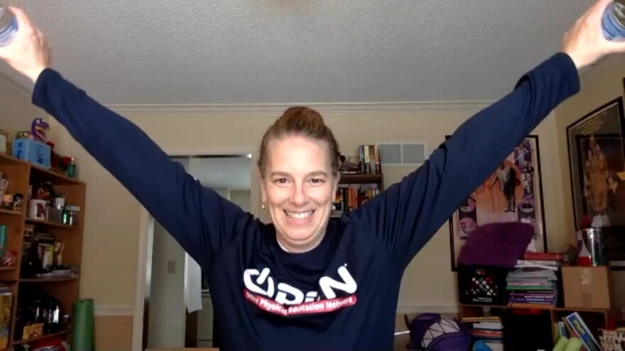 Gym teacher Stephanie Dickson leads a remote class from her home office. Here, she's doing a butterfly motion using cans of food for hand weights.