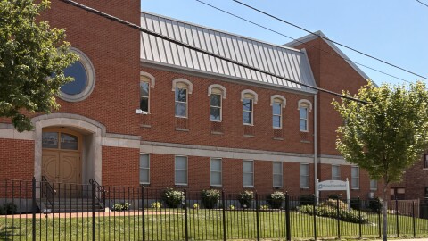 A large brick building with a grassy lawn in front and a black fence separating it from the sidewalk.