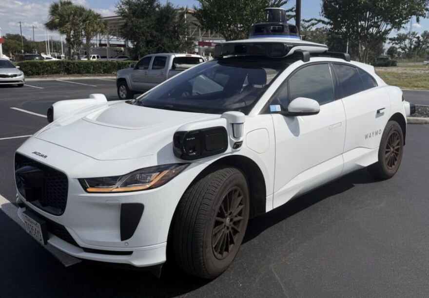 A white Jaguar car with black sensors on the front sides and on the roof sits in a parking lot in-between spots.