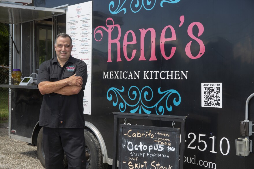 A man in a black button up shirt and black pants stands with his arms crossed in front of a food truck that says Rene's Mexican Kitchen in pink, white and blue.