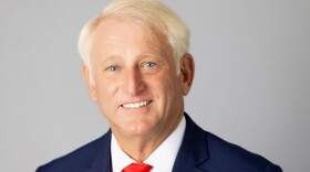 A headshot of a man with white hair, blue eyes, a blue shirt and a red tie