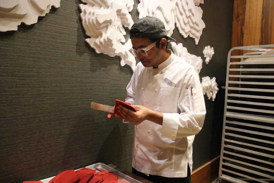A man in a backwards baseball cap, glasses and a white chef coat holds a red piece of gingerbread and a rectangular grater. He is shaving one end of the red gingerbread piece.