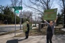 People stand along the side of a road holding up signs. One reads "Save the Park." Another: "Congestion Cut-Throughs Traffic Jams"