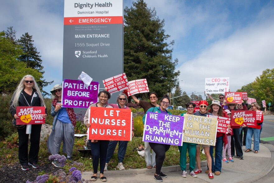 A group of people hold signs that say things like, "let nurses nurse," and "we keep the department running," stand on a street corner outside the hospital. 