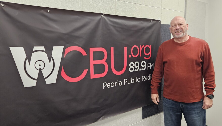 A man in a red sweater stands next to a WCBU 89.9 FM Peoria Public Radio banner on a wall.