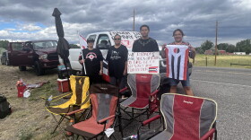 Four people hold protest signs and flags in front of a truck with more signs on it. Lawn chairs are set up in front of them.