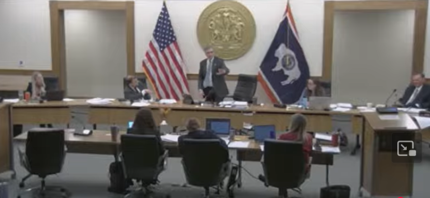 Five people sit at a long desk. The man in the center is standing and gesturing with his left hand.