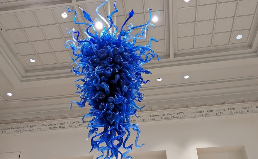 A blue, glass sculpture hangs in the air inside an open, white space.