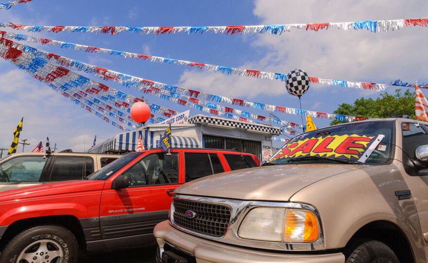 A vibrant car dealership scene with several vehicles parked in front. Colorful banners and flags, including red, silver, and blue streamers, are strung overhead. A prominent "SALE" sign is displayed on the windshield of a beige SUV. In the background, there is a small building adorned with a racing-themed checkered pattern.