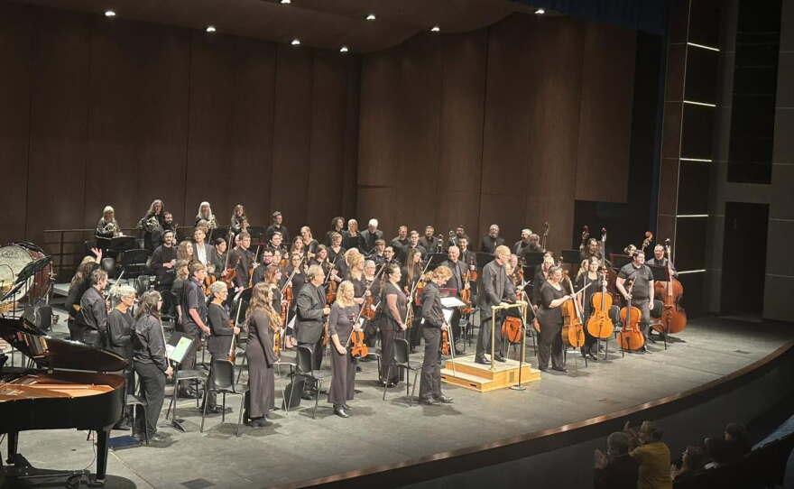 An orchestra all wearing black facing the audience on a grey stage.