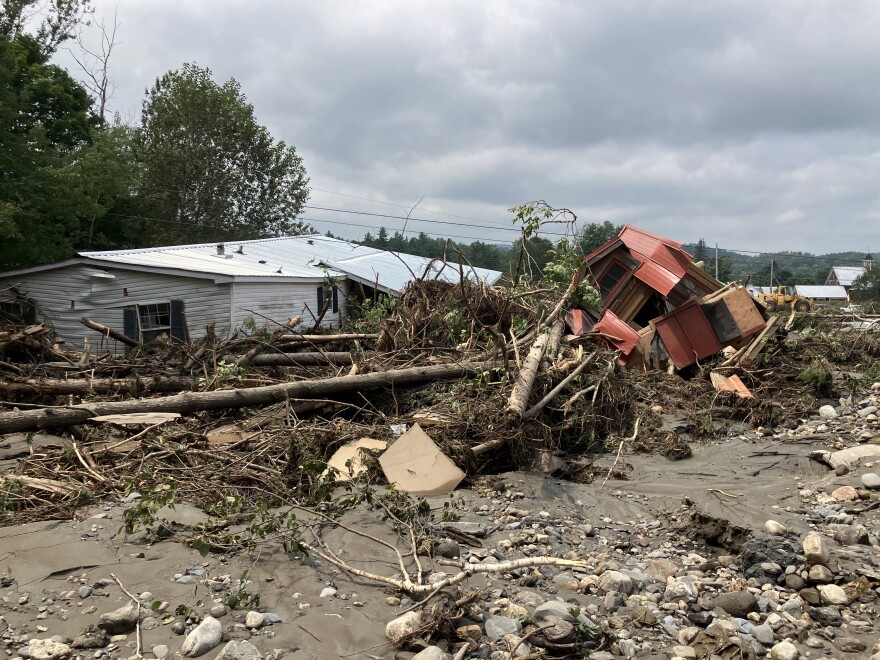 A damaged home and a damaged tiny home are part of a pile of downed trees and rubble.