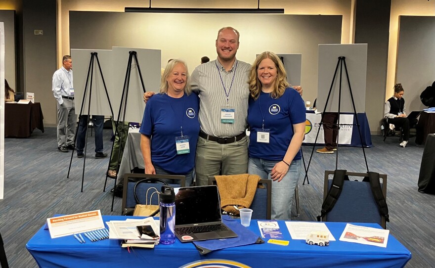 Three people stand in front of a table at a conference. The table reads Deer River Full Service Schools.