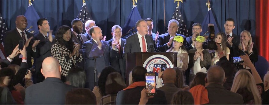 Lee Zeldin at a podium surrounded by supporters, with U.S. and state flags behind them and an audience in front of them.