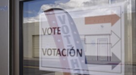 Una ventana con un cartel que dice: «Vote, Votoción», con varias flechas apuntando a la derecha. En el reflejo hay otro cartel de votación vertical que dice: «Vote here».