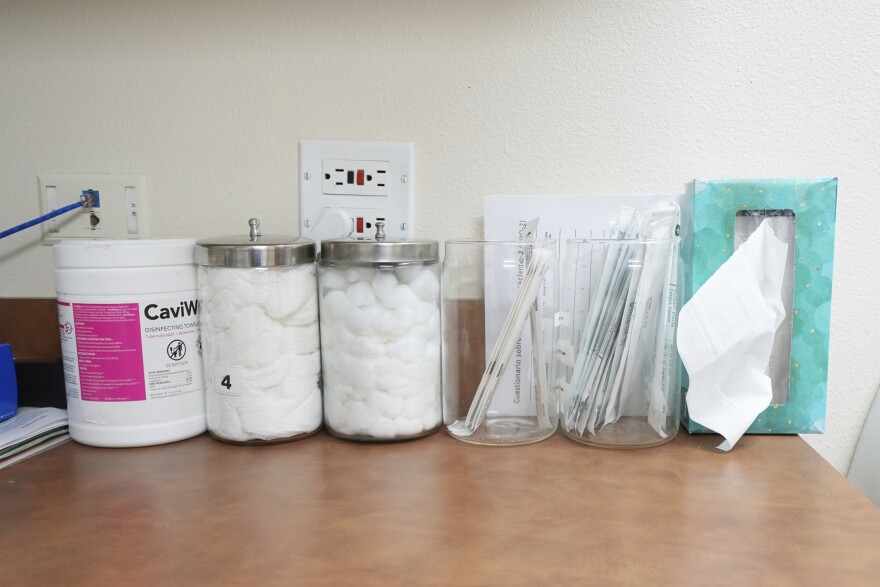 Cotton balls in jars, a box of tissues set with the bottom against the all, and other medical supplies are seen on a brown countertop