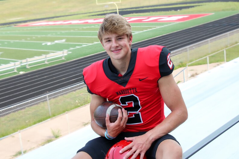 A young man with blonde hair smiles into the camera. He’s wearing a red football jersey, with black lettering. The jersey says “Marquette.” The jersey number is 2. He sits on a bleacher, holding a football, with the football field behind him.