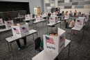 Photo of a large room where there are many tables with cardboard "Vote" dividers set up on them People are walking around and sitting at the tables filling out ballots.