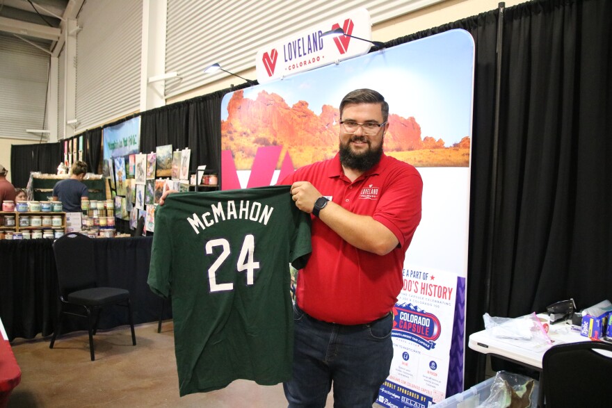 A man in a red shirt at a booth holds up a green shirt that reads "McMahon 24" in white letters. 