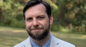 A young man with brown hair and beard wearing a white suit, blue pinstriped shirt and dark blue tie, smiling at the camera