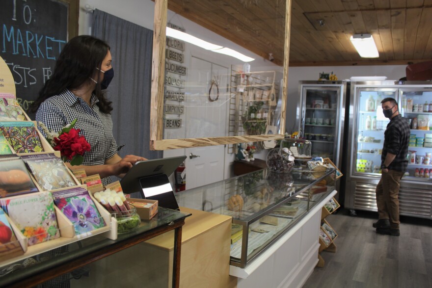 A woman wearing a dress and a mask stands behind a Plexiglas shield at a cash register in a farm store. A man, to the right, looks in a cooler.