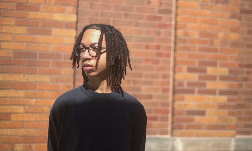 A young man with glasses stands in the sunlight in front of a brick wall.