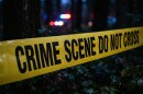 Yellow crime scene tape with the message "Crime Scene Do Not Cross" blocks a wooded area with police cruiser lights in the background