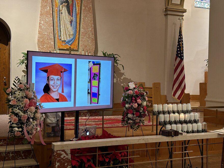 A digital screen showing an artist's picture of a young girl in a red cap and gown stands next to flowers and candles at the front of a church. 