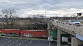 The Franklin Street bridge today, with modern vehicles crossing above active railroad tracks, a passing train below, and a parking lot with semitrailers.