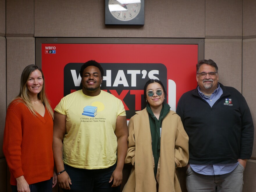 A woman in red, a man in yellow, a woman in a tan jacket, and a man in a black sweater stand in front of a red, black, and white What's Next? sign