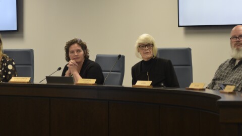 Three women sit with one man on the right. All are seated behind a desk, and looking at a speaker not in frame.