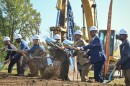 Men and women wearing business attire and wearing white hard hats used shiny, silver shoves to toss dirt in front of yellow earth-moving equipment