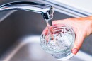 A photo shows a woman's hand holding a glass of clear, clean drinking water under a tap.
