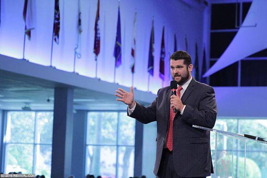 A bearded white man wearing a blue suit and red tie holds microphone on a stage